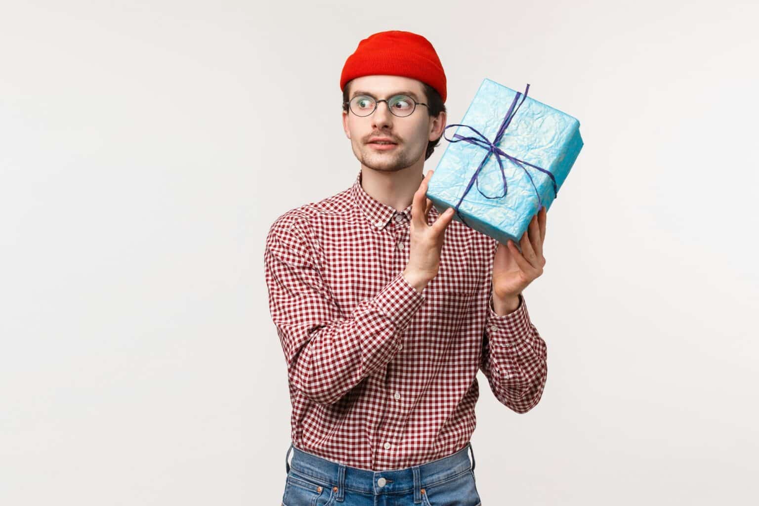 Waist-up portrait excited caucasian bearded guy in red beanie, shaking blue wrapped gift box near