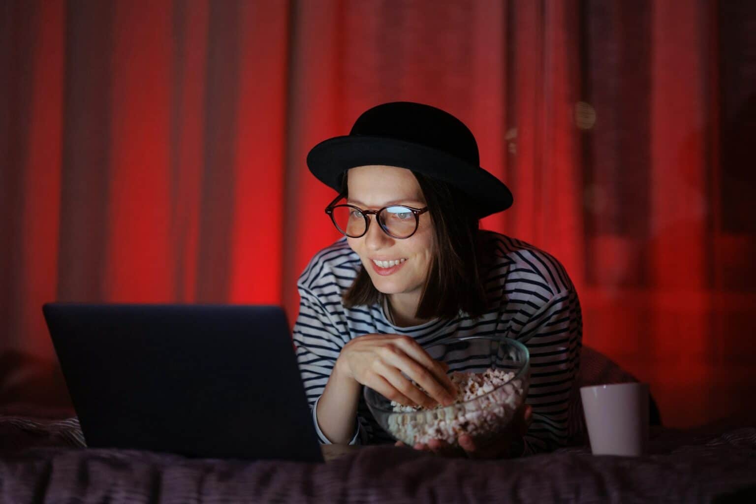 woman in a dark room with a hat at home watching TV or a movie on a laptop and eating popcorn.
