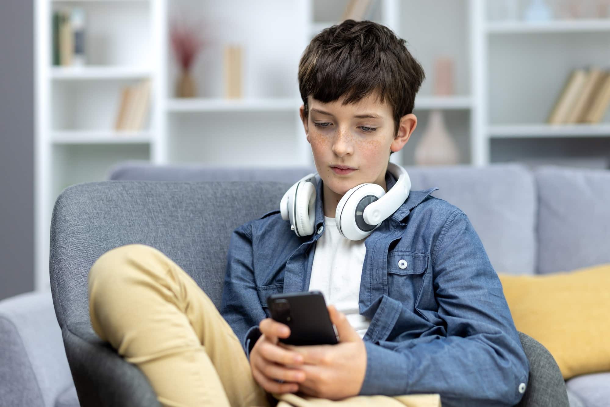 Serious child of a teenage boy in headphones sitting at home on the sofa, using the phone. Listens
