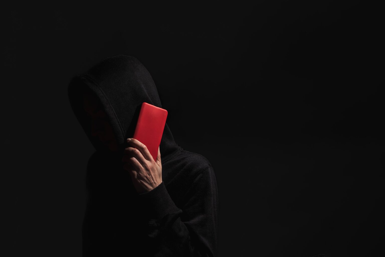 Hacker with hidden face holding red smartphone on dark background,internet security and cyberattacks